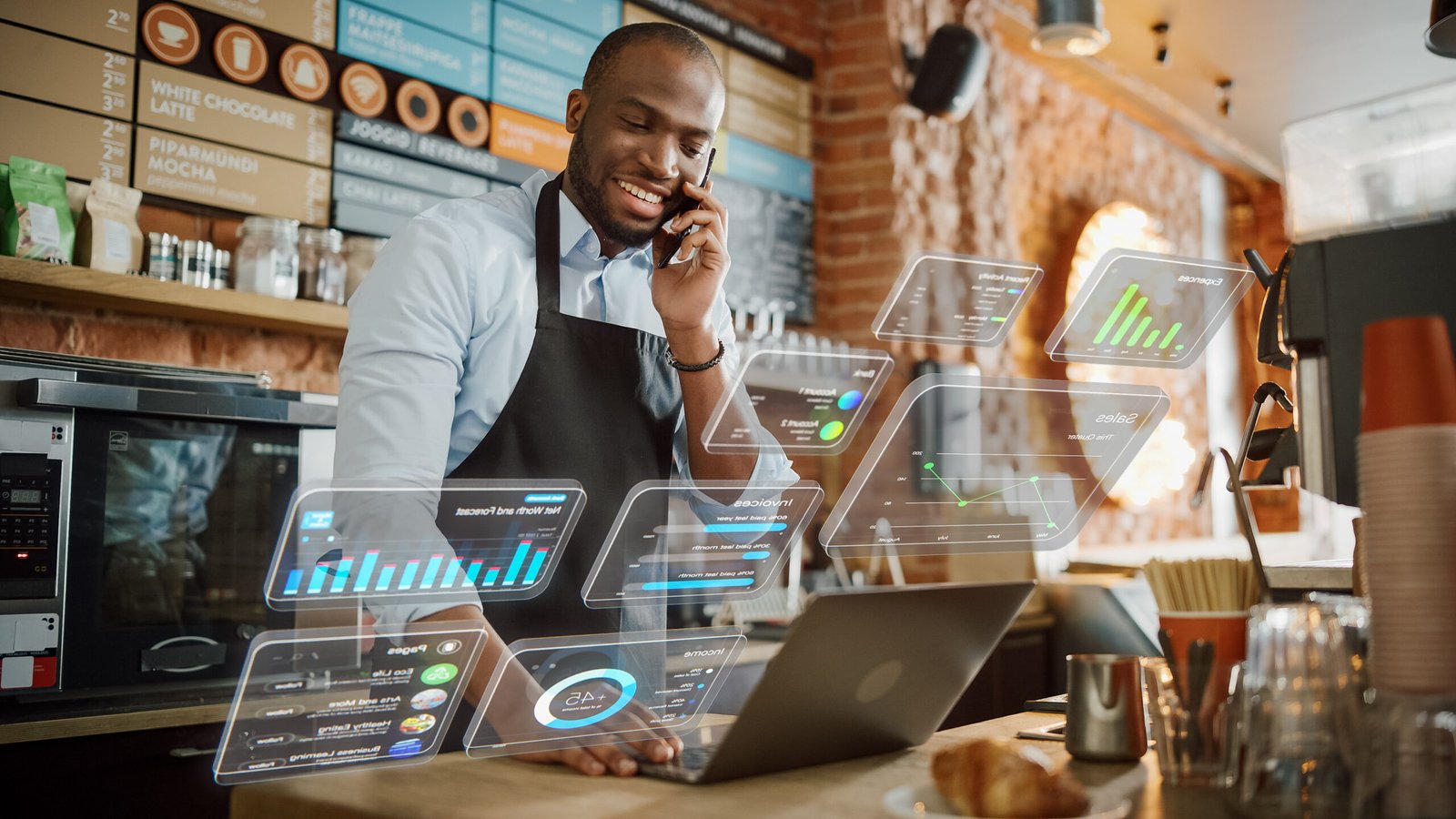 African American Coffee Shop Owner is Working on Computer and Talking on a Phone in a Cozy Cafe. Augmented Reality Icons Popping Out of Restaurant Manager's Laptop: Small Black Business Concept.